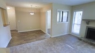 This single-floor space presents a clean, functional layout with a seamless transition between a carpeted living area and an adjoining dining or flex space finished with wood-look hardwood flooring. The two distinct flooring materials create a natural visual division between the zones while maintaining an open, connected feel throughout.

The living area features neutral-toned carpet, contributing to a warm and comfortable atmosphere, while the adjacent dining area showcases rich, wood-toned planks that add a touch of elegance and are easy to maintain. Warm beige walls run throughout, providing a neutral backdrop that complements both flooring choices and offers versatility for various décor styles.

A pendant light fixture hangs above the dining area, serving as a subtle focal point and providing dedicated overhead illumination for that zone. Multiple windows along the walls invite an abundance of natural light, brightening the interior and creating an airy, welcoming ambiance. White window blinds offer privacy control while maintaining the clean aesthetic. An interior doorway visible from the main living space suggests additional rooms or a hallway beyond, hinting at a well-organized floor plan.