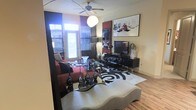 This single-floor space showcases a bold and contemporary interior design aesthetic that immediately captures attention. The living area serves as the centerpiece, featuring warm hardwood flooring that grounds the room and contrasts elegantly with the dramatic black-and-white color palette throughout. A large, intricately patterned area rug anchors the seating arrangement, where a rich red sofa adds a vibrant pop of color against the otherwise monochromatic scheme. The room is furnished with a sleek glass coffee table adorned with sculptural black vase accents, alongside a modern entertainment console housing a flat-screen television. A striking piece of wall art depicting a chess-themed scene adds an intellectual and artistic focal point to the space. Natural light flows generously through the windows, which are dressed with bold geometric patterned curtains that complement the overall design theme. A ceiling fan with an integrated light fixture enhances both comfort and illumination. The overall ambiance is sophisticated and expressive, blending dramatic design choices with functional living spaces to create an interior that feels curated, dynamic, and full of personality.