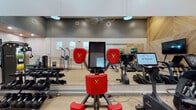 Located at 3600 University Drive in Durham, NC, this contemporary fitness center offers a well-appointed, single-floor layout designed to support a comprehensive range of workout routines. The space features soaring ceilings that contribute to an open, airy atmosphere, with clerestory windows along the upper walls allowing natural light to filter into the main exercise area.

The cardio zone is lined with a full row of True-brand treadmills, ellipticals, stair climbers, and Expresso interactive cycling machines, all positioned along a floor-to-ceiling mirrored wall that amplifies the sense of space. A wood-paneled accent wall adds a warm, modern aesthetic contrast to the sleek dark ceiling.

The strength and functional training area features a dedicated TRX suspension training rig, a robust rack of kettlebells and resistance tools, medicine balls, BOSU balance trainers, and True-brand selectorized weight machines. A wooden ballet barre with cable railing delineates this functional zone. A separate enclosed studio area with glass doors is visible, providing a quieter space for focused training or classes.

Free weights, including a full dumbbell rack, round out the equipment offerings. The light wood-toned flooring, recessed and pendant lighting, and cohesive design elements throughout give the facility a polished, professional appearance suited for serious fitness pursuits.