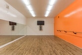 Located at 1050 Summerside Dr in San Jose, CA, this single-room dance studio is purpose-built for movement and instruction. The open floor plan offers generous, unobstructed space ideal for classes, rehearsals, or personal practice. The standout feature is a full-length mirrored wall that spans the width of the studio, allowing dancers to monitor their form and technique with ease. Complementing the mirrors are wall-mounted ballet barres that run along multiple walls, including the bold orange accent wall that energizes the space with warmth and visual contrast against the clean white walls and ceiling. The studio's polished hardwood floor provides the ideal surface for dance, offering both durability and a smooth, responsive feel underfoot. Overhead, rows of bright fluorescent lighting ensure the entire space is evenly and thoroughly illuminated. A wall-mounted flat-screen TV accompanied by decorative geometric shelving provides a functional media station for instructional use or music playback. The space is also equipped with visible HVAC venting and ductwork to maintain a comfortable environment during active use. Compact yet efficient, this well-appointed studio provides everything needed for a fully functional dance and movement facility.