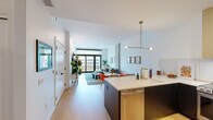 Located at 3515 Delgany St in Denver's vibrant RiNo neighborhood, this modern 1-bedroom, 2-bathroom residence offers a thoughtfully designed open-concept layout with contemporary finishes throughout. Light-toned hardwood flooring flows through the main living area, connecting the kitchen, dining space, and living room in a seamless arrangement. The kitchen is outfitted with dark cabinetry extending to ceiling height, stainless steel appliances including a French door refrigerator and electric range, and a central island with a white countertop and gold-tone fixtures. A sculptural brass pendant light anchors the kitchen-dining transition. The living room is bright and airy, featuring large floor-to-ceiling sliding glass doors that open to a private balcony with city views, complemented by an exposed concrete column that adds an industrial character. The bedroom offers carpeted floors, large windows with urban views, a ceiling fan, and direct access to a walk-in closet. The primary bathroom is a standout, showcasing blush-pink wall tile, a floating double vanity in warm wood tones, white countertops, and gold-accented fixtures and mirrors. A second bathroom provides additional convenience. Clean white walls and recessed lighting throughout give the space a bright, modern ambiance.