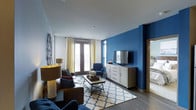 This single-floor living space makes a strong visual impression through a carefully considered interior design scheme. The living room serves as the centerpiece, anchored by a striking deep blue accent wall that contrasts sharply with the surrounding white walls and ceiling, creating a dynamic and modern aesthetic. Dark hardwood-style flooring runs throughout, providing a warm, grounding element beneath the space.

Natural light flows generously into the room through large windows and a set of French doors dressed with yellow and patterned curtains, adding a touch of softness and warmth to the bold color palette. The doors appear to open to an outdoor area, enhancing the sense of openness.

The living area is thoughtfully furnished with a mix of a brown leather sofa, navy blue armchairs, and a geometric patterned area rug that ties the color palette together. A rectangular coffee table anchors the seating arrangement. A white multi-drawer media console houses a wall-mounted flat-screen television against the blue feature wall. Decorative accents, including a circular wall ornament and a table lamp, complete the curated, welcoming ambiance of the space.