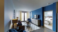 This single-floor living space makes a strong visual impression through a carefully considered interior design scheme. The living room serves as the centerpiece, anchored by a striking deep blue accent wall that contrasts sharply with the surrounding white walls and ceiling, creating a dynamic and modern aesthetic. Dark hardwood-style flooring runs throughout, providing a warm, grounding element beneath the space.

Natural light flows generously into the room through large windows and a set of French doors dressed with yellow and patterned curtains, adding a touch of softness and warmth to the bold color palette. The doors appear to open to an outdoor area, enhancing the sense of openness.

The living area is thoughtfully furnished with a mix of a brown leather sofa, navy blue armchairs, and a geometric patterned area rug that ties the color palette together. A rectangular coffee table anchors the seating arrangement. A white multi-drawer media console houses a wall-mounted flat-screen television against the blue feature wall. Decorative accents, including a circular wall ornament and a table lamp, complete the curated, welcoming ambiance of the space.