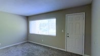 This single-story space features a main living area finished with wall-to-wall carpet in a neutral tone, providing a comfortable and understated foundation. The room is characterized by its wide, horizontal window fitted with white horizontal blinds, which allows a generous amount of natural light to fill the space while offering views of the surrounding greenery outdoors. The walls are painted in a soft, neutral beige-gray tone, contributing to a calm and versatile ambiance. A six-panel white entry door is positioned adjacent to the window, providing direct access and adding a clean, classic architectural detail to the room. White baseboards frame the perimeter of the room, offering a crisp contrast against the carpeted floor and painted walls. A textured ceiling completes the interior finish, consistent with standard residential construction. The overall layout is open and unobstructed, making it adaptable for a variety of furnishing arrangements. The combination of neutral finishes, natural light, and outdoor visibility gives the space a simple, functional, and welcoming character suitable for everyday living.
