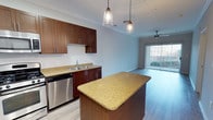 This single-floor residence offers a thoughtfully designed open-concept layout that seamlessly connects the kitchen and living areas. The kitchen is a standout feature, showcasing rich dark wood cabinetry, granite-look countertops, and a central island that provides additional prep space and casual seating. Stainless steel appliances, including a dishwasher, complement the modern aesthetic, while a double-basin sink is framed beneath upper cabinetry for practical functionality. Stylish pendant lights hang above the island, adding warmth and a contemporary touch to the space. Crown molding along the ceiling adds a refined architectural detail throughout. The open living area extends naturally from the kitchen, featuring light wood-style flooring that runs throughout and a ceiling fan that enhances comfort. Large sliding glass doors at the far end flood the space with natural light and provide direct access to an outdoor area, offering a seamless indoor-outdoor connection. The overall ambiance is bright, clean, and inviting, combining practical design with modern finishes to create a comfortable and functional living environment.
