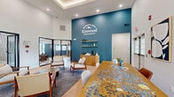 The Kinwood Apartments community clubhouse at 2 River Chase in Rensselaer, NY, is a thoughtfully designed single-floor amenity space offering residents a broad range of gathering and wellness areas. The welcoming lobby greets visitors with soaring ceilings, light hardwood flooring, a classic fireplace, and bold teal accent walls bearing the Kinwood Apartments branding. Large glass doors and windows invite abundant natural light throughout.

Adjacent to the entry, a comfortable lounge area features warm leather sofas, mid-century modern seating, and curated artwork set against two-tone teal walls, creating an inviting atmosphere for relaxation or socializing. A café-style dining nook with tufted leather banquette seating and green-topped tables provides an intimate spot for casual gatherings.

A vibrant media and game room showcases a custom powder-blue built-in entertainment unit with shelving, a large wall-mounted TV, colorful artwork, and playful furnishings, including rounded ottomans and light wood accent tables. Natural light pours in through large windows.

The expansive fitness center is a standout feature, boasting a distinctive green ceiling, rubber flooring, floor-to-ceiling mirrors, and a comprehensive selection of equipment including treadmills, ellipticals, cable machines, free weights, and strength-training stations, all supported by ample natural light from multiple windows.