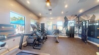 This fitness center is thoughtfully designed to offer a comprehensive workout experience within a single-floor layout. The main exercise area is dominated by a bold, artistic wall mural depicting a dramatic mountain landscape, creating an energizing and visually striking backdrop for workouts. The space is outfitted with a robust selection of Matrix-brand equipment, including multiple treadmills, elliptical machines, a stair climber, and a multi-function cable strength training station, catering to a wide range of fitness routines.

Durable rubber flooring throughout the main workout area provides both comfort and safety underfoot. Multiple windows line the walls, flooding the space with natural light and offering views of a resort-style outdoor pool area, complete with palm trees and vibrant blue umbrellas. Recessed lighting and a ceiling fan complement the natural illumination.

A dedicated stretch and flexibility area is separated by elegant white-framed glass French doors and features blue accent walls, large wall mirrors, and a shelving unit stocked with yoga mats, foam rollers, and fresh towels. This area is finished with carpet flooring and opens through the glass doors into an adjacent, warmly lit lounge space with wood flooring, beamed ceilings, and a decorative chandelier — creating a seamless transition between active and relaxation zones.