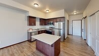 This single-floor, one-bedroom, one-bathroom unit offers a thoughtfully designed layout that maximizes its compact footprint. The open-concept kitchen and living area forms the heart of the space, featuring rich dark-stained wood cabinetry, light-toned countertops with a subtle tile backsplash, and a full suite of stainless steel appliances including a range, microwave, dishwasher, and refrigerator. A central kitchen island with a white countertop provides additional workspace and casual seating. Light wood-style flooring runs consistently throughout, unifying the space and adding warmth.

The living area is generously proportioned relative to the overall footprint, offering an open, airy feel with neutral walls and room to configure furniture comfortably. The bedroom receives ample natural light through a large window overlooking greenery, contributing a bright and tranquil atmosphere. Neutral tones and clean lines keep the room feeling open despite its cozy dimensions.

The bathroom is notably functional, incorporating a stacked in-unit washer and dryer alongside a dark wood vanity, toilet, and a towel bar, maximizing practicality within the space. Consistent flooring and bright lighting maintain the cohesive aesthetic found throughout the unit.