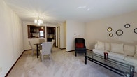 This single-floor space offers a well-organized, open layout that seamlessly connects the living, dining, and kitchen areas. The living area is anchored by plush, light-toned carpeting and features a white sofa paired with a dark accent chair adorned with a bold red pillow, creating a classic contrast. A rectangular glass-top coffee table adds a contemporary touch, while the neutral cream walls contribute to a clean, inviting ambiance throughout.

The dining area sits adjacent to the living space and accommodates a glass-top dining table surrounded by upholstered chairs, ideal for everyday meals or casual gatherings. A crystal chandelier overhead serves as a refined focal point, lending an elegant detail to the shared open space.

Beyond the dining area, the kitchen is visible through an open passthrough, showcasing rich, dark wood cabinetry, stainless steel appliances, and a window that allows natural light to filter in. The dark cabinetry creates a striking contrast against the lighter tones of the surrounding spaces. Dark wood baseboards frame the entire layout, adding definition and a finished look throughout this well-appointed, single-level space.