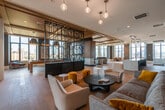This upscale amenity floor is a masterclass in contemporary design, offering a seamless blend of functionality and refined aesthetics. The entry corridor sets an immediate tone with marble-veined wall panels, geometric patterned wallcoverings, polished tile flooring, and elegant circular pendant lighting that guides residents into the expansive interior.

The open-plan layout is anchored by wide-plank hardwood flooring that flows throughout, unifying the various zones. A dramatic decorative steel screen with bold geometric patterns acts as a defining architectural partition, delineating spaces while maintaining visual connectivity. Warm wood-panel ceilings add depth and texture overhead.

The lounge area features a large wall-mounted display framed in rich wood paneling above a linear fireplace, surrounded by carefully curated seating and floor-to-ceiling windows that frame sweeping city skyline views. A wet bar with stone countertops, a stainless steel sink, glass-front cabinetry, and a beverage cooler provides a well-appointed hub for entertaining.

A separate dining area benefits from dramatic oversized circular pendant lights and additional panoramic windows, while the open social lounge incorporates vibrant, contemporary furnishings accented by curated artwork and sculptural wall installations. Large glass doors lead to an outdoor terrace, extending the living space and inviting abundant natural light deep into the interior.