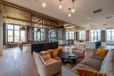 This upscale amenity floor is a masterclass in contemporary design, offering a seamless blend of functionality and refined aesthetics. The entry corridor sets an immediate tone with marble-veined wall panels, geometric patterned wallcoverings, polished tile flooring, and elegant circular pendant lighting that guides residents into the expansive interior.

The open-plan layout is anchored by wide-plank hardwood flooring that flows throughout, unifying the various zones. A dramatic decorative steel screen with bold geometric patterns acts as a defining architectural partition, delineating spaces while maintaining visual connectivity. Warm wood-panel ceilings add depth and texture overhead.

The lounge area features a large wall-mounted display framed in rich wood paneling above a linear fireplace, surrounded by carefully curated seating and floor-to-ceiling windows that frame sweeping city skyline views. A wet bar with stone countertops, a stainless steel sink, glass-front cabinetry, and a beverage cooler provides a well-appointed hub for entertaining.

A separate dining area benefits from dramatic oversized circular pendant lights and additional panoramic windows, while the open social lounge incorporates vibrant, contemporary furnishings accented by curated artwork and sculptural wall installations. Large glass doors lead to an outdoor terrace, extending the living space and inviting abundant natural light deep into the interior.