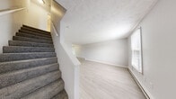 This two-floor residence offers a practical and functional layout across three bedrooms and one bathroom. The main floor hosts a combined kitchen and living room area, where light wood-look flooring creates a clean, bright atmosphere. The kitchen features white upper cabinetry, gray lower cabinets, marble-style countertops, and black appliances including a full-size refrigerator, electric range, and dishwasher — all compactly arranged for efficient use of space. The adjacent living area is finished with the same light flooring, a textured ceiling, and a baseboard heating system, with a window that brings in natural light and a view of the outdoors. A carpeted staircase with a white handrail connects the main floor to the upper level, where all three bedrooms are located. The primary bedroom offers a comfortable footprint, a window with natural light, and baseboard heating. The two additional bedrooms are finished with neutral carpet, light gray walls, and clean white trim. The single full bathroom includes a soaking bathtub with a tiled surround. Throughout the home, a neutral color palette and consistent finishes give the space a cohesive, move-in-ready feel.
