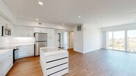 Located at 1011-1013 Everett St in the Echo Park neighborhood of Los Angeles, this modern 2-bedroom residence offers a clean, contemporary design across a single floor. The open-concept layout seamlessly connects the kitchen and living room, creating a bright and functional shared space. The kitchen is a standout feature, outfitted with sleek white cabinetry extending to the ceiling, a full suite of stainless steel appliances including a refrigerator, range, microwave, and dishwasher, and a central island with a white countertop and built-in storage drawers. Light wood flooring flows throughout the entire unit, complementing the crisp white walls and recessed lighting. The living area is bathed in natural light, with large sliding glass doors opening to a balcony that offers expansive views of the surrounding Los Angeles skyline and lush greenery. The primary bedroom is generously proportioned and includes a built-in wardrobe with warm-toned hardware and corner windows that frame panoramic city views. The second bedroom also benefits from natural light and includes a closet with built-in shelving. The overall ambiance is modern, minimalist, and airy, with a consistent design language carried throughout each room.