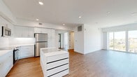 Located at 1011-1013 Everett St in the Echo Park neighborhood of Los Angeles, this modern 2-bedroom residence offers a clean, contemporary design across a single floor. The open-concept layout seamlessly connects the kitchen and living room, creating a bright and functional shared space. The kitchen is a standout feature, outfitted with sleek white cabinetry extending to the ceiling, a full suite of stainless steel appliances including a refrigerator, range, microwave, and dishwasher, and a central island with a white countertop and built-in storage drawers. Light wood flooring flows throughout the entire unit, complementing the crisp white walls and recessed lighting. The living area is bathed in natural light, with large sliding glass doors opening to a balcony that offers expansive views of the surrounding Los Angeles skyline and lush greenery. The primary bedroom is generously proportioned and includes a built-in wardrobe with warm-toned hardware and corner windows that frame panoramic city views. The second bedroom also benefits from natural light and includes a closet with built-in shelving. The overall ambiance is modern, minimalist, and airy, with a consistent design language carried throughout each room.