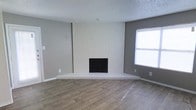 This single-story space presents a clean, neutral aesthetic well-suited for a variety of uses. The living area is anchored by a striking white-painted brick fireplace, which extends from floor to ceiling and commands attention as the room's defining architectural feature. The fireplace is fitted with classic black double doors and sits atop a raised brick hearth, adding dimension and visual weight to the space.

The flooring throughout consists of wood-style planks in a warm, medium tone, providing a cohesive and durable surface that contrasts nicely with the cool gray wall color. The walls are painted in a soft, medium gray, lending the space a modern and understated character. A textured popcorn ceiling adds a traditional touch overhead.

Natural light enters through a window positioned to the right of the fireplace, brightening the space and highlighting the clean lines of the interior. Electrical outlets are conveniently distributed along the baseboards, and the space appears freshly updated and move-in ready. Overall, the room offers a comfortable and functional layout with a timeless, neutral palette.
