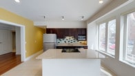This single-floor space showcases a thoughtfully designed open-concept kitchen that serves as the heart of the layout. The kitchen is appointed with rich, dark espresso cabinetry that provides generous storage, complemented by a sleek mosaic tile backsplash in neutral tones with contrasting dark accents. A spacious granite-topped island anchors the space, featuring an integrated cooktop and offering ample room for meal preparation or casual dining.

Stainless steel appliances, including a full-sized refrigerator and built-in microwave, add a contemporary, professional-grade feel to the kitchen. A dedicated sink with a modern faucet is set into the countertop along the back wall, enhancing functionality. Light tile flooring runs throughout, lending a clean and cohesive look to the space.

A bold yellow accent wall introduces a warm, energetic pop of color that contrasts beautifully with the white walls and ceilings. Recessed and track lighting provide excellent illumination throughout. A large window floods the kitchen with natural light, creating a bright and inviting atmosphere. Overall, the space combines modern finishes with a practical, open layout that is both functional and visually appealing.