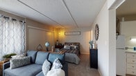 This single-floor unit offers an efficient and thoughtfully arranged open-concept layout that seamlessly combines living and sleeping areas within one cohesive space. The main room is carpeted throughout and features a textured ceiling, neutral wall tones, and a large window dressed with patterned sheer curtains that allow natural light to fill the room. A wall-unit air conditioner is positioned beneath the window, providing climate control directly within the living space.

The open layout accommodates both a sectional sofa and a full-sized bed, clearly defining separate living and sleeping zones without physical division. A sliding-door closet is situated along one wall of the sleeping area, offering convenient built-in storage. The living area includes space for a television console and area rug, contributing to a comfortable, organized atmosphere.

The kitchen is a separate, enclosed galley-style space featuring white cabinetry extending to the ceiling, a drop-in sink with light-colored countertops, and an electric range. Wood-look flooring runs through the kitchen, contrasting with the carpeted main area.

The bathroom includes a tub-shower combination with a curtain rod, a toilet, and a vanity with speckled countertops, complemented by wood-look flooring. The overall ambiance of the unit is practical and inviting, with a cohesive neutral color palette throughout.