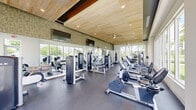 Located in Homestead, FL, this fitness center offers a thoughtfully designed exercise space that combines modern aesthetics with comprehensive workout functionality. The room features a distinctive slatted wood ceiling with recessed lighting and a ceiling fan, creating a warm, contemporary atmosphere enhanced by crisp white walls accented with textured green wall panels.

Large floor-to-ceiling windows line multiple walls, flooding the space with abundant natural light and offering views of the surrounding tropical landscaping, including palm trees and lush greenery. The rubber-floored workout area is well-organized and equipped for a full range of training modalities. Cardio equipment includes treadmills, elliptical machines, and stationary bikes, while a dedicated strength zone houses Cable machines and multi-use weight stations. A separate area is designated for functional and combat training, featuring heavy punching bags, medicine balls, and a flat bench for free weight exercises. Full-length mirrors along one wall enhance the sense of openness and allow users to monitor their form. Wall-mounted televisions are positioned throughout the space for added convenience. The overall ambiance is bright, open, and inviting, making this fitness center a well-rounded facility suited for a variety of workout routines.
