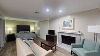 This single-story studio suite offers a thoughtfully designed open-concept layout that efficiently combines living, sleeping, and dining functions within one cohesive space. Hardwood-style flooring runs throughout, lending a warm, consistent aesthetic across all areas.

The main living area serves as the focal point of the suite, anchored by a tile-surround fireplace with a white mantel and framed artwork above. A flat-screen television on a dark wood dresser, a sofa, armchair with teal accents, and a coffee table complete the comfortable seating arrangement. Vertical blinds along one wall provide privacy and light control.

The sleeping area is integrated seamlessly into the open layout, featuring a queen bed with a dark upholstered headboard, flanked by nightstands with lamps. A mirrored closet offers convenient storage nearby, along with a full-size refrigerator tucked into an alcove.

The kitchen is a functional galley-style space equipped with dark cabinetry, granite countertops, stainless steel appliances including an electric range and microwave, a stainless sink with a flexible faucet, and a white subway tile backsplash.

The bathroom features a dark vanity with granite countertops, a large mirror, and hardwood-style flooring, maintaining the suite's cohesive design throughout.