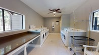 This shared laundry facility is a well-maintained, single-floor space designed for practical, everyday use. The room is finished with neutral gray walls and light-toned flooring, creating a clean and bright environment. Multiple front-loading washers and dryers are arranged along both sides of the room, providing ample capacity for residents. A centrally mounted ceiling fan with an integrated light fixture ensures proper air circulation and comfortable conditions throughout the space. A built-in folding counter with a wood-toned surface runs along one side of the room, offering a convenient area for sorting and folding laundry. A wall-mounted payment kiosk is positioned for easy access, streamlining the process of operating the machines. Informational postings are displayed near the machines to guide users. The space also accommodates a laundry cart for added convenience. A window allows natural light to filter into the room, enhancing visibility. The layout is open and uncluttered, allowing multiple users to move freely and comfortably. Overall, this laundry room is a functional and well-organized shared amenity that prioritizes efficiency and ease of use.