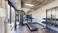 This fitness center delivers a functional and visually dynamic workout environment defined by its industrial-inspired architecture and lively interior design. The exposed concrete ceiling with visible piping and overhead fluorescent lighting gives the space a modern, urban character, while durable wood-look flooring runs throughout, providing a clean and practical surface underfoot.

A bold, large-scale botanical mural featuring monstera leaves and tropical foliage covers one full wall, adding vibrant color and an energizing aesthetic to the space. Floor-to-ceiling glass along one side of the room allows ample natural light to pour in and provides views to the outdoors, keeping the interior feeling open and airy.

The fitness center is thoroughly equipped for a complete workout. Cardio offerings include a treadmill and a stationary cycling bike, while a multi-station cable machine supports a wide variety of strength exercises. A full rack of dumbbells in graduated weights, an adjustable workout bench, and a wall-mounted flat-screen television round out the amenities. A large mirror along one wall visually expands the space and is useful for monitoring form during exercise.

The layout is efficient and well-organized, making effective use of the available floor space to accommodate multiple users simultaneously. The combination of natural light, industrial design elements, and vibrant wall art creates an inviting and motivating atmosphere for daily fitness routines.