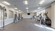 This fitness center is a single-floor facility designed to support a full range of workout routines. The space is laid out openly, allowing for comfortable movement between equipment zones. Flooring transitions between carpet and rubber-textured surfaces, providing appropriate traction and cushioning throughout the room.

The cardio section lines one side of the facility and is outfitted with multiple treadmills, elliptical trainers, and a stationary bike, all positioned to take advantage of the large windows that line the exterior wall, allowing generous natural light and outdoor views to enhance the workout experience.

The strength training area features a cable machine, a leg press, and additional resistance machines, all arranged along a mirrored wall that amplifies the sense of space and supports proper form during exercise. A multi-tiered dumbbell rack offers a broad range of free weights for supplemental training, accompanied by a weight bench.

A wall-mounted flat-screen television is centrally positioned for visibility across the room. The drop-panel ceiling is fitted with recessed fluorescent lighting that ensures consistent, even illumination throughout the space. A clearly posted fitness area rules board and direct exterior door access add to the facility's practical, user-friendly design. The overall ambiance is clean, modern, and functional.