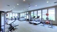 This modern fitness center is designed to support a comprehensive workout experience in a bright, open environment. The space is lined with large windows along multiple walls, allowing abundant natural light to fill the room and offering views of the surrounding greenery, creating an energizing atmosphere for exercise. The flooring is a light-toned hardwood, contributing to the clean and contemporary aesthetic throughout.

The cardio section is prominently featured, with a row of treadmills positioned to take advantage of the natural light and outdoor views. Additional cardio equipment includes ellipticals and stationary bikes, providing a variety of aerobic workout options. A multi-functional cable and strength machine is centrally positioned, offering users versatile resistance training capabilities. Free weight benches are also available for targeted strength training.

Ceiling fans and recessed lighting are evenly distributed across the ceiling, ensuring comfortable airflow and consistent illumination throughout all hours of use. The overall layout is thoughtfully organized, with ample space between equipment to allow for safe and comfortable movement. The neutral gray walls and clean design elements give the space a professional and welcoming ambiance suited for fitness enthusiasts of all levels.