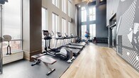 Located at 518 W Fayette St in Baltimore, MD, this fitness center is a thoughtfully designed exercise space that combines functionality with striking architectural character. The room is defined by soaring double-height ceilings and expansive floor-to-ceiling windows along multiple walls, flooding the interior with abundant natural light and offering impressive panoramic views of the Baltimore skyline.

The flooring transitions between warm light wood and durable rubber mat zones, clearly delineating cardio and strength training areas. A row of Life Fitness treadmills lines the windowed wall, complemented by elliptical trainers and a stair climber — all positioned to take full advantage of the sweeping city views during workouts. The strength training section features a multi-station cable machine, stationary and spin bikes, a rowing machine, and a well-stocked dumbbell rack with a full range of free weights and resistance equipment.

An elegant sputnik-style chandelier overhead adds a refined design touch, contrasting beautifully with the deep charcoal wall tones that lend the space a modern, upscale aesthetic. The combination of premium equipment, dramatic natural light, and polished design details makes this fitness center a standout amenity in the heart of downtown Baltimore.