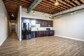 This single-floor urban loft showcases a compelling blend of industrial character and modern comfort. The defining architectural feature is the dramatic exposed wood beam ceiling, which spans the open-concept living and kitchen area, complemented by visible metal ductwork and pendant lighting that reinforce the industrial aesthetic. Light wood-tone luxury vinyl flooring runs throughout the main living space, reflecting natural light that pours in through a set of large windows along one wall.

The kitchen is thoughtfully designed with rich, dark-stained wood cabinetry extending from floor to ceiling, light countertops, a stainless steel sink, and a full suite of black appliances including a refrigerator, range, microwave, and dishwasher. The open layout of the main area provides generous, flexible living space.

The bedrooms offer a quieter, more intimate atmosphere, transitioning to plush carpeted flooring and neutral gray walls. Exposed structural steel columns and ductwork carry the industrial theme into these rooms as well. The bathroom is clean and well-appointed, featuring large-format tile flooring, a full bathtub with shower, a dark wood vanity with white countertop, and recessed lighting for a polished finish.