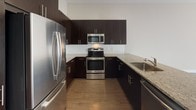 This thoughtfully designed single-floor residence offers a cohesive blend of modern finishes and functional layout throughout. The kitchen is a standout feature, showcasing rich dark cabinetry, granite countertops, a classic white tile backsplash, hardwood flooring, and a full suite of stainless steel appliances including a refrigerator, range, microwave, and dishwasher. A peninsula counter provides additional prep space and opens toward the adjoining living and dining area.

The living area is bathed in natural light through expansive floor-to-ceiling sliding glass doors and large windows that frame outdoor views of the surrounding streetscape and greenery, creating an airy, open ambiance. Smooth hardwood floors extend through this space, complementing the neutral wall tones.

The bedrooms are carpeted in plush neutral tones, offering comfort and warmth. Each bedroom features generous closet space with standard rod and shelf configurations, as well as windows fitted with roller shades for light control and privacy.

Two bathrooms serve the residence. The full bathroom includes a soaking tub with an overhead rain showerhead, white tile surround, dark vanity cabinetry with granite countertops, and light tile flooring. A second, more compact bathroom features a similarly styled dark vanity with granite countertop and matching tile flooring, providing convenient access throughout the unit.
