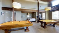 This single-floor recreation space is designed for entertainment and social engagement. The layout is open and expansive, anchored by two full-size pool tables — one in a classic honey-toned wood finish and another in a darker walnut-style finish — each positioned beneath large, modern drum pendant lights that provide focused, even illumination. The high ceilings, accented by prominent rustic wooden beams and posts, lend the space a warm, lodge-like architectural character that contrasts stylishly with the neutral sage and beige wall tones. Carpeted flooring runs throughout, adding comfort underfoot. Toward the rear of the space, a casual lounge area features a white sofa and soft ambient lighting, offering a place to relax between games. An arcade game cabinet is visible along the far wall, further enhancing the recreational atmosphere. Natural light filters in through a window near the arcade area. Wall art and decorative pendants contribute subtle personality throughout the space. Overall, the combination of rustic structural elements, generous ceiling height, and thoughtfully arranged entertainment amenities creates a welcoming and well-appointed recreational environment.