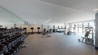 This expansive fitness center is thoughtfully designed across a single level, offering distinct zones tailored to a comprehensive workout experience. The main cardio area is generously proportioned, housing a lineup of treadmills, ellipticals, stationary bikes, and a stair climber. Floor-to-ceiling mirrors line one wall, amplifying the sense of space, while large glass doors open to an outdoor courtyard, flooding the area with natural light and providing views of the surrounding buildings.

Adjacent to the cardio zone, a dedicated strength training section features a multi-station cable machine, an assortment of free weights on organized racks, adjustable benches, and stability balls, all set against darker gray walls that give this area a focused, energetic atmosphere. Linear LED lighting above adds a sleek, modern touch throughout both zones.

A separate studio space rounds out the facility, featuring rich hardwood flooring, wall-mounted ballet barres, and full mirrored walls that reflect natural light streaming in through large glazed panels. This versatile studio is well-suited for yoga, barre, dance, or group fitness classes. The overall design is clean and contemporary, combining neutral tones, durable flooring materials, and ample open floor space to create an inviting and functional fitness environment.