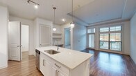 Located at 2925 Buford Dr in Buford, GA, this single-floor unit offers a clean, modern aesthetic highlighted by an open-concept layout and thoughtfully selected finishes throughout. The kitchen serves as the centerpiece of the main living area, anchored by a large island with a quartz countertop, an undermount sink, and pendant lighting overhead. White shaker-style cabinetry, a hexagonal tile backsplash, and a full suite of stainless steel appliances—including a range, over-the-range microwave, and French door refrigerator—complete the space. Track lighting and recessed fixtures provide ample illumination. The kitchen flows seamlessly into the living area, which features warm hardwood-style flooring, crown molding, a tray ceiling, and a glass door that opens to a private balcony. Built-in shelving and a built-in desk add functional detail to this shared living space. The bedrooms are carpeted for comfort and receive natural light through large windows; at least one bedroom includes a walk-in closet. The bathroom is finished with a white vanity, quartz countertop, chrome fixtures, and a wood-framed mirror, maintaining the unit's cohesive, contemporary style throughout.