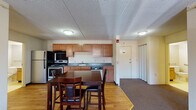 This single-floor residential unit offers a practical and functional layout suited for everyday living. The kitchen is well-appointed with warm wood cabinetry, dark countertops, stainless steel appliances including a refrigerator, electric range, and dishwasher, all set atop durable hardwood flooring. A dining area adjacent to the kitchen comfortably accommodates a table and chairs, creating a natural gathering space. The transition from kitchen to living areas is marked by a shift to plush carpeting, which continues throughout the living room and bedrooms. The living room features a large window that allows natural light to filter in, offering views of surrounding greenery. Two generously sized bedrooms each include ceiling fans, carpeted floors, baseboard heating, and windows with curtains. Both bathrooms are designed with accessibility in mind, featuring grab bars, vinyl tile flooring, wood vanities, and ample space for ease of movement. One bathroom includes a walk-in shower enclosure. The unit's overall design emphasizes practicality and accessibility, with a neutral color palette and functional finishes throughout, making it a well-rounded and livable space.