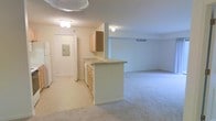 This single-floor residential unit offers a practical and open layout that seamlessly connects the kitchen and living areas. The kitchen is outfitted with light wood cabinetry, a built-in sink, and a peninsula counter that extends into the main living space, providing both additional workspace and a natural dividing point between the two areas. White appliances, including a refrigerator, complement the neutral cabinetry and light-toned countertops. The flooring throughout is plush wall-to-wall carpeting in a neutral beige tone, adding warmth and continuity across the space. The living area is generously proportioned, offering ample room for a variety of furniture arrangements. Overhead flush-mount light fixtures provide consistent illumination throughout. The hallway visible near the kitchen leads to additional rooms, with a paneled door and an electrical panel accessible nearby. A ceiling vent indicates a central HVAC system for climate control. The overall color palette is kept light and neutral — creamy whites and soft beiges — lending the space a clean, understated aesthetic that is move-in ready and easily personalized.