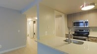 This single-floor space showcases a thoughtfully designed open-concept layout with a seamless flow between living and functional areas. The kitchen is equipped with stainless steel appliances, including a range and built-in microwave, and features light wood cabinetry paired with a speckled quartz peninsula countertop and a contemporary coil-neck faucet with an undermount sink. The peninsula creates a natural division between the kitchen and the adjoining living area while maintaining an open, airy feel throughout.

Light-toned hardwood-style flooring runs consistently across the space, contributing to a bright and cohesive aesthetic. Walls are finished in a clean, neutral palette that enhances the sense of openness. A dedicated in-unit laundry area is conveniently tucked within a hallway corridor, housing a stacked washer and dryer behind accessible cabinetry. The hallway leads further to what appears to be a bathroom, reinforcing the efficient and practical layout of the space. Recessed and flush-mount ceiling lighting provides consistent illumination throughout.