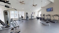 This fitness center offers a spacious, well-organized workout environment designed to accommodate a range of exercise needs. The room features vaulted ceilings that lend an open, airy feel, enhanced by multiple ceiling fans that ensure consistent air circulation throughout the space. Recessed lighting complements the abundant natural light streaming in through several large windows fitted with white blinds, creating a bright and inviting atmosphere.

The durable rubber flooring provides both safety and comfort underfoot, making it suitable for high-impact workouts. The facility is outfitted with a comprehensive selection of equipment, including strength-training machines such as cable and resistance units, as well as cardio equipment including elliptical trainers and a rowing machine. The machines are thoughtfully arranged to allow for comfortable movement between stations.

The walls are finished in a clean, neutral white, contributing to the room's open and motivating ambiance. A piece of artwork adds a subtle decorative touch. Overall, the space is designed to be functional, comfortable, and conducive to an effective fitness routine.