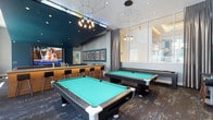 This dynamic single-floor social lounge is designed for entertainment and community gathering, offering a well-appointed space that blends modern aesthetics with recreational functionality. At the heart of the room, two full-sized pool tables with striking teal felt take center stage, set atop a richly patterned area rug that anchors the space with a sense of depth and style. Overhead, an eye-catching array of geometric wire pendant lights and clustered Edison-style fixtures cast a warm, ambient glow across the room, complementing the space's contemporary design language. A built-in bar counter with sleek bar stools lines one wall, providing a casual seating area ideal for socializing, while a large wall-mounted display screen serves as a focal point for viewing entertainment. The walls feature a tasteful mix of teal and neutral gray tones, adorned with framed artwork that adds a curated, gallery-like touch. Floor-to-ceiling glass panels along one side of the room invite natural light and offer a visual connection to adjacent spaces. The overall ambiance is energetic yet refined, making this lounge a standout communal amenity suited for both casual relaxation and lively social events.