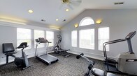 Located at 4426 Calderwood Drive in Columbus, Ohio, this single-floor fitness facility offers a well-appointed workout space designed to support an active lifestyle. The exercise room is the centerpiece of the space, boasting soaring vaulted ceilings that lend an open, airy feel to the environment. A ceiling fan keeps the room comfortable during workouts, while recessed lighting and wall-mounted sconces provide ample illumination throughout. Natural light floods the room through an abundance of windows, including a striking arched window that serves as a distinctive architectural focal point. White blinds on each window allow for flexible light control. The space is outfitted with a variety of fitness equipment, including a treadmill, an elliptical cross-trainer, and a multi-function strength training machine, making it suitable for both cardiovascular and resistance training. Durable, textured carpet flooring covers the workout area, providing cushioning and stability underfoot. The neutral gray walls and clean architectural lines create a calm, focused atmosphere conducive to exercise. Overall, this fitness space combines thoughtful design with practical functionality.