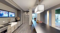 This single-floor space showcases a refined contemporary aesthetic, seamlessly blending functionality with upscale design. The interior is defined by a cohesive grey color palette complemented by warm gold hardware and decorative accents, creating an atmosphere of understated elegance.

The space features rich wood-look flooring in a deep charcoal tone, adding warmth and texture underfoot while contrasting beautifully against the crisp white and grey cabinetry. Built-in cabinetry with brass hardware lines one wall, providing generous storage while contributing to the polished, tailored character of the space.

A long, dark countertop bar area serves as a central gathering feature, accompanied by slender gold-toned bar stools that reinforce the sophisticated design language. Above, a striking geometric pendant light with a latticed shade makes a bold architectural statement while providing focused illumination.

Decorative plates arranged artfully on the wall and metallic accent pieces add curated visual interest throughout. A wall-mounted television is integrated seamlessly into the built-ins. Natural light filters in through a window at the far end, offering views of a lush outdoor setting and brightening the overall ambiance of this well-appointed space.