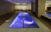 Located at 75 Sterling Blvd in Englewood, NJ, this sophisticated indoor aquatic and relaxation facility is thoughtfully designed to offer a serene, spa-like environment. The centerpiece of the space is a generously sized indoor lap pool, its luminous blue water illuminated from within, creating a calming and inviting atmosphere throughout. Flanking the pool are stacked-stone accent columns and accent walls that lend a warm, earthy texture, contrasting beautifully with the clean white walls and contemporary recessed lighting overhead. The pool deck is finished with large-format wood-look tile flooring, adding warmth and continuity across the space. An ADA-compliant pool lift is installed poolside, ensuring accessibility for all users. A dedicated lounge area lines one side of the pool, furnished with natural teak slatted chaise lounges and matching side tables, providing a comfortable space for rest and relaxation. Pendant lighting and wall sconces contribute a soft, ambient glow, enhancing the spa-like feel. A wooden shelving unit offers convenient storage within the lounge area. The layout also includes an adjacent wet area with a handheld shower fixture and a vessel sink. The overall design strikes a refined balance between natural materials and modern finishes, creating a welcoming and functional wellness environment.