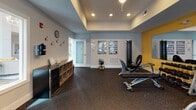 Located at 1001 Scotney Ln in Myrtle Beach, SC, this single-floor fitness center is thoughtfully designed to accommodate a wide range of workout styles and fitness levels. The space is divided into distinct functional zones, each outfitted with professional-grade equipment. The cardio area features multiple treadmills and elliptical machines positioned along a row of windows, allowing natural light to fill the room and creating an open, energizing atmosphere. A tray ceiling with recessed lighting and a ceiling fan adds architectural detail while maintaining comfort during workouts. The strength training zone is lined with large wall mirrors and accented by bold yellow walls, creating a dynamic and motivating environment. It houses a cable machine, a recumbent bike, and additional weight training equipment. A dedicated functional training area includes wall-mounted storage for stability balls in multiple sizes, foam rollers, a medicine ball rack, and step platforms, all kept neatly organized. The entry area provides coat hooks, a beverage cooler, open cubbies for personal belongings, and informational fitness charts for reference. Throughout the space, durable rubber flooring ensures safety and supports high-impact activity. The overall design balances practicality with an inviting aesthetic, making it a well-rounded facility for daily fitness routines.