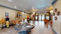 The Arbour Valley clubhouse and leasing office is a thoughtfully designed single-floor community space that combines functionality with warm, inviting aesthetics. The main lobby greets visitors with large-format tile flooring, recessed lighting, and a striking round pedestal table at its center, serving as a natural focal point. French doors flanked by draped windows flood the entry with natural light, creating an open and airy atmosphere. The Arbour Valley branded wall feature anchors the seating area, where clusters of patterned and solid upholstered chairs invite relaxed conversation around a glass-top coffee table. A dedicated library nook offers a large glass-door bookcase stocked with reading materials, complemented by a wall-mounted television and decorative shelving. The kitchen is well-appointed with dark espresso cabinetry, granite-style countertops, a double basin sink, a full-size refrigerator, and a breakfast bar with barstool seating—ideal for community gatherings or staff use. A private office enclosed by French doors with glass panes provides a quiet workspace. Crown molding, recessed and pendant lighting, and cohesive warm-toned décor throughout give the space a polished, welcoming character suited for both residents and staff.