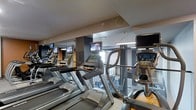 This modern fitness center is thoughtfully designed to provide a comprehensive workout environment within a single-level space. The gym is outfitted with a row of Matrix-brand treadmills and elliptical machines, each equipped with individual entertainment screens, allowing users to stay engaged during cardio sessions. The layout is organized and spacious, offering ample room to move between equipment comfortably.

The interior features a clean, contemporary aesthetic with a neutral color palette of grays and whites, accented by sleek structural columns and recessed overhead lighting that keeps the space bright and energizing. A large mirror spans one wall, enhancing the sense of openness and allowing users to monitor their form during workouts. The mirrored wall also reflects a weight and resistance training area visible in the background, suggesting the facility extends beyond cardio equipment to include a broader range of fitness options.

The flooring appears to be a durable, light-toned surface suitable for high-traffic athletic use. Pendant lighting fixtures add a polished touch to the overall design. The space strikes a balance between functional gym utility and modern interior design, making it a welcoming and well-appointed facility for daily fitness routines.