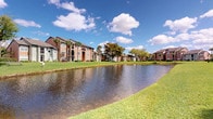 This single-floor residence is nestled within a beautifully landscaped multi-building community that immediately impresses with its tranquil outdoor setting. The community grounds are defined by a picturesque pond that reflects the surrounding greenery and open sky, creating a peaceful natural focal point. Expansive, well-manicured lawns stretch along the water's edge, bordered by mature palm trees and leafy deciduous trees that lend the property a lush, tropical character consistent with a warm Florida climate. The residential buildings feature a traditional architectural style with warm brick and earth-toned exteriors, accented by pitched rooflines in deep red and grey tones. Balconies and windows face outward toward the scenic grounds, offering residents pleasant views of the water and landscaping. The community's layout encourages an open, airy atmosphere with generous space between structures, promoting both privacy and a sense of community. The combination of natural water features, mature vegetation, and classic building design gives the property a welcoming and well-established character that is both functional and visually appealing.