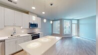 Located at 2008–2032 N Beauregard St in Alexandria, VA, this single-floor residence presents a thoughtfully designed, modern interior with a clean, neutral aesthetic throughout. Light hardwood-style flooring runs continuously across the main living areas, lending warmth and visual cohesion to the space.

The kitchen is a standout feature, offering a generous island with a bright white countertop, flat-front white cabinetry extending to the ceiling, and a full suite of stainless steel appliances including a refrigerator, range, microwave, and dishwasher. A classic subway tile backsplash and recessed pendant lighting above the island complete the contemporary look. The kitchen flows seamlessly into an open living area bathed in natural light from large windows with horizontal blinds, offering views of the surrounding neighborhood.

A distinctive sliding barn door with frosted glass panels provides stylish access to a spacious walk-in closet, adding a modern architectural detail to the bedroom area. The bathroom features light wood-tone flooring, a tiled tub-shower combination, a white vanity with an undermount sink, and clean, minimal finishes throughout. The overall ambiance of the space is bright, airy, and move-in ready.
