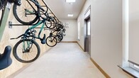 Located at 19007 E 59th Ave in Denver, Colorado, this dedicated bike storage room offers a practical and well-organized space designed specifically for bicycle storage. The room is configured as a hallway-style layout, providing a clear, open floor area that allows for easy movement and bike retrieval.

The standout feature of this space is its wall-mounted vertical bike rack system, installed along a wood-paneled accent wall. The racks efficiently maximize storage capacity by suspending multiple bicycles — including road bikes and mountain bikes — off the floor, keeping the central area unobstructed and accessible.

The room is finished with neutral-toned walls and warm wood baseboards, giving it a clean and functional aesthetic. Overhead, a flush skylight panel provides natural light, supplemented by recessed ceiling lighting to ensure the space remains well-lit at all times. The concrete floor is durable and easy to maintain, well-suited for the wear associated with bicycle use.

A wall-mounted heater unit ensures the space remains climate-appropriate year-round. A bike storage policy sign is posted near the entry door, offering clear guidelines for users. Natural light also enters through a window fitted with a roller shade. The exterior-facing door with a window provides secure, convenient access directly from outside, making this room a highly functional amenity for cyclists.