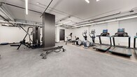 Located at 200 Elgin St in the heart of Ottawa, this fitness facility offers a generously sized, single-floor exercise room designed to support a wide range of workout routines. The space features a clean, modern aesthetic with crisp white walls, dark baseboards, and durable rubberized flooring throughout, creating a functional and professional environment for physical training.

The exercise room is thoughtfully organized into distinct activity zones. A dedicated strength training area houses a multi-functional cable machine with integrated weight stacks, a tiered dumbbell rack stocked with a full range of free weights, and an adjustable workout bench — catering to both compound and isolated exercises. A separate cardio zone features a lineup of modern equipment including treadmills, elliptical trainers, and a stationary bike, each equipped with digital displays. Additional open floor space accommodates balance equipment, resistance tools, and folding exercise mats, ideal for stretching or functional training.

Overhead, exposed industrial-style ductwork and piping run across the ceiling alongside bright linear fluorescent lighting, giving the facility a practical, no-frills atmosphere. Interior windows allow visual connectivity between sections of the space. The layout is efficient and open, ensuring ample room to move freely between stations, making this a comprehensive and accessible fitness facility.