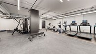 Located at 200 Elgin St in the heart of Ottawa, this fitness facility offers a generously sized, single-floor exercise room designed to support a wide range of workout routines. The space features a clean, modern aesthetic with crisp white walls, dark baseboards, and durable rubberized flooring throughout, creating a functional and professional environment for physical training.

The exercise room is thoughtfully organized into distinct activity zones. A dedicated strength training area houses a multi-functional cable machine with integrated weight stacks, a tiered dumbbell rack stocked with a full range of free weights, and an adjustable workout bench — catering to both compound and isolated exercises. A separate cardio zone features a lineup of modern equipment including treadmills, elliptical trainers, and a stationary bike, each equipped with digital displays. Additional open floor space accommodates balance equipment, resistance tools, and folding exercise mats, ideal for stretching or functional training.

Overhead, exposed industrial-style ductwork and piping run across the ceiling alongside bright linear fluorescent lighting, giving the facility a practical, no-frills atmosphere. Interior windows allow visual connectivity between sections of the space. The layout is efficient and open, ensuring ample room to move freely between stations, making this a comprehensive and accessible fitness facility.