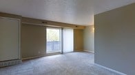 This single-floor residence offers a clean and functional layout designed for comfortable everyday living. The main living area is a notable highlight, featuring soft, wall-to-wall carpeting that adds warmth underfoot and a textured ceiling that gives the space a classic, finished look. Neutral-toned walls in warm beige and muted green provide a versatile backdrop suitable for a variety of interior styles.

Natural light fills the living room through a large window and a sliding glass door, which opens onto a private balcony with views of surrounding greenery. Sheer vertical blinds dress the door and window, allowing for both light diffusion and privacy control. Ceiling-mounted air vents indicate a central HVAC system for year-round climate comfort. A wall-mounted light fixture provides ambient illumination, complementing the natural light sources.

The open feel of the living space, combined with the outdoor access, creates a welcoming and airy environment. The single-floor layout ensures easy accessibility and seamless flow throughout the unit. Overall, this space presents a straightforward, well-maintained residential setting with practical features and a calm, understated aesthetic suited to a range of lifestyles.